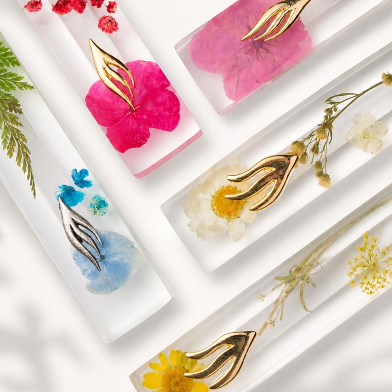 K&ouml;nnte beinhalten: Nahaufnahme mehrerer rechteckiger Harzst&uuml;cke, die jeweils eine andere getrocknete Blume enthalten. Die Blumen sind in verschiedenen Farben, darunter Rosa, Gelb und Wei&szlig;. Jedes St&uuml;ck hat einen dekorativen Metallakzent.
