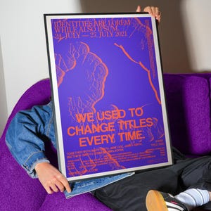 Pu&ograve; includere: Un poster incorniciato con sfondo viola e testo arancione. Il poster recita "IDENTITIES ARE LOREM WHILE ALSO IPSUM. 24 LUGLIO - 27 LUGLIO 2021" e "WE USED TO CHANGE TITLES EVERY TIME." Il poster &egrave; tenuto da una persona.