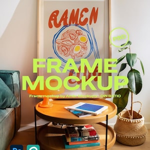 Op de afbeelding: Een ingelijste poster met het woord "RAMEN" in oranje letters. De poster toont een kom ramen met eieren en eetstokjes. Een houten bijzettafel met twee niveaus bevat boeken en een lamp. Rechts staat een plant in een geweven mand.