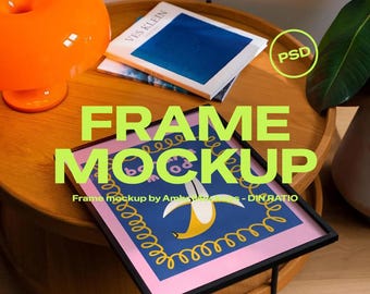 May include: A framed art print with a banana design, displayed on a wooden table. The artwork features a black frame, a pink border, and a blue background with a yellow banana illustration. An orange lamp and books are also on the table.