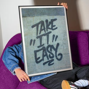 Pu&ograve; includere: Una stampa artistica incorniciata con le parole "TAKE IT EASY" in lettering nero in stile graffiti. La stampa &egrave; tenuta da una persona con una giacca di jeans, seduta su un divano viola. L'opera d'arte ha uno sfondo grigio strutturato.