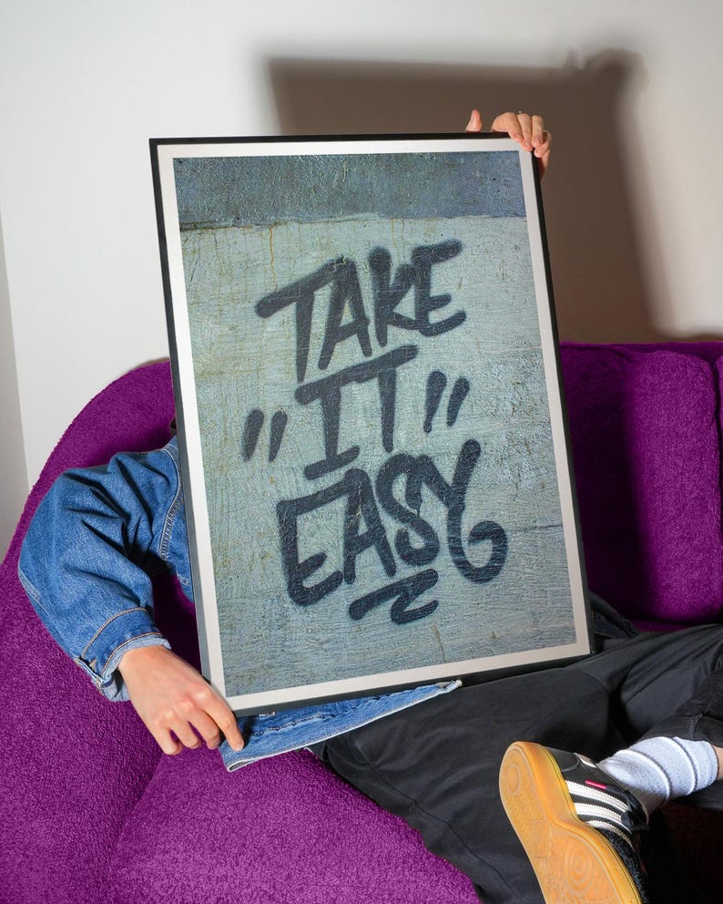 Pu&ograve; includere: Una stampa artistica incorniciata con le parole "TAKE IT EASY" in lettering nero in stile graffiti. La stampa &egrave; tenuta da una persona con una giacca di jeans, seduta su un divano viola. L'opera d'arte ha uno sfondo grigio strutturato.