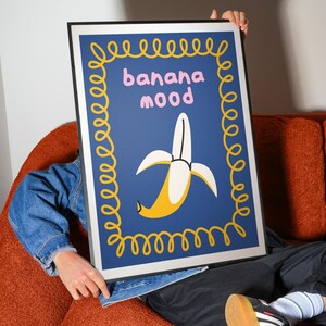 Pu&ograve; includere: Una stampa artistica incorniciata con un design a banana stilizzato. L'opera d'arte ha uno sfondo blu, un bordo giallo e le parole "banana mood" in rosa. La banana &egrave; gialla e bianca.