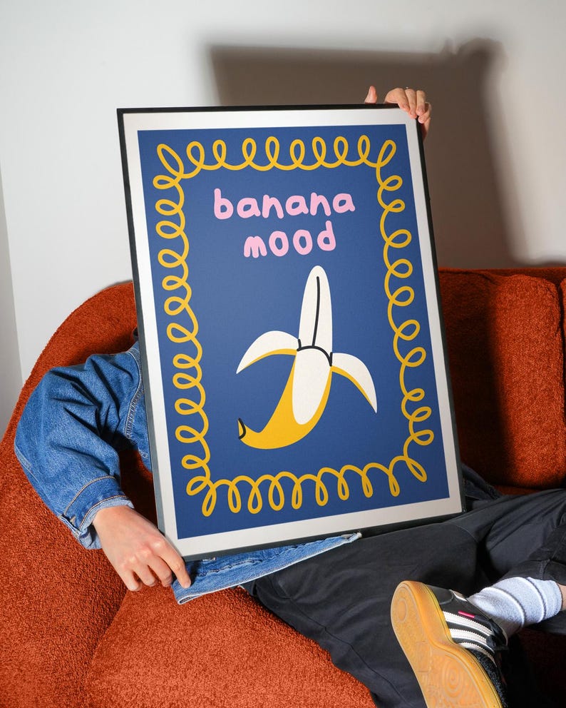 Pu&ograve; includere: Una stampa artistica incorniciata con un design a banana stilizzato. L'opera d'arte ha uno sfondo blu, un bordo giallo e le parole "banana mood" in rosa. La banana &egrave; gialla e bianca.