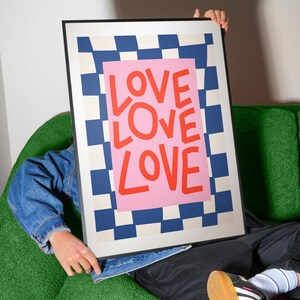 Pu&ograve; includere: Una stampa artistica incorniciata con la parola "LOVE" in grandi lettere rosse su sfondo rosa. Lo sfondo presenta un motivo a scacchiera blu e bianco. La stampa &egrave; tenuta da una persona.
