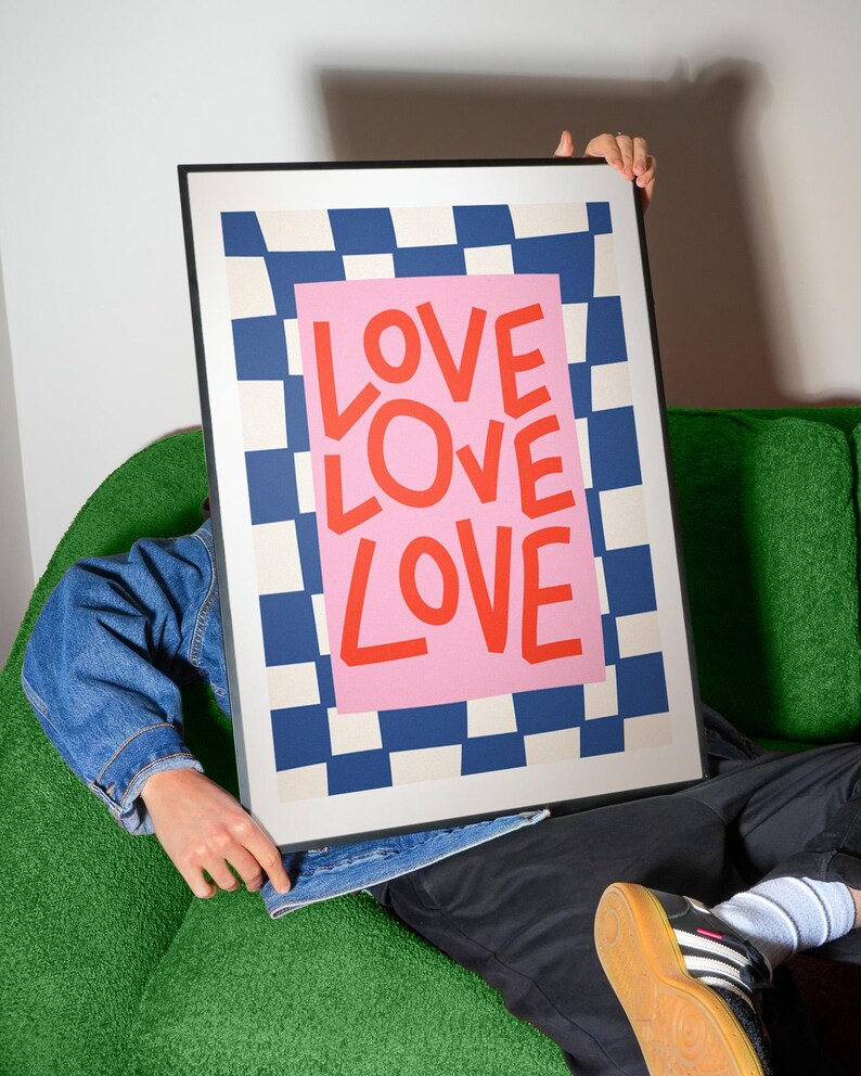 Pu&ograve; includere: Una stampa artistica incorniciata con la parola "LOVE" in grandi lettere rosse su sfondo rosa. Lo sfondo presenta un motivo a scacchiera blu e bianco. La stampa &egrave; tenuta da una persona.