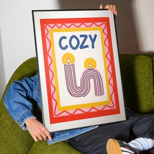 Pu&ograve; includere: Una stampa artistica incorniciata con la parola "COZY" in lettere blu. La stampa presenta due candele stilizzate con strisce rosa e nere e fiamme gialle, su uno sfondo bianco con bordi rossi, rosa e gialli. La cornice &egrave; nera.