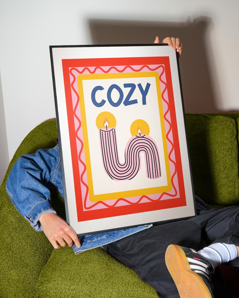 Pu&ograve; includere: Una stampa artistica incorniciata con la parola "COZY" in lettere blu. La stampa presenta due candele stilizzate con strisce rosa e nere e fiamme gialle, su uno sfondo bianco con bordi rossi, rosa e gialli. La cornice &egrave; nera.