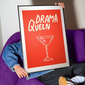 Pu&ograve; includere: Un poster rosso incorniciato con le parole "DRAMA QUEEN" in scrittura bianca e un'illustrazione di cocktail. Il poster &egrave; tenuto davanti a una persona seduta su un divano viola. La persona indossa una giacca di jeans e pantaloni neri.