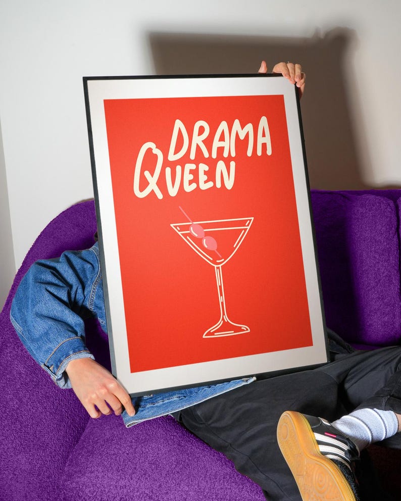 Pu&ograve; includere: Un poster rosso incorniciato con le parole "DRAMA QUEEN" in scrittura bianca e un'illustrazione di cocktail. Il poster &egrave; tenuto davanti a una persona seduta su un divano viola. La persona indossa una giacca di jeans e pantaloni neri.