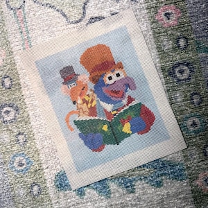 May include: A needlepoint canvas featuring a colorful design of a blue creature with a large nose wearing a brown top hat and a small tan rat wearing a grey top hat, both characters holding an open green book against a light blue background.