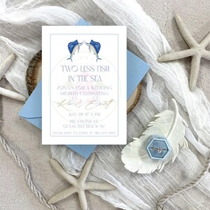 Può includere: Un invito per un bridal shower con il testo "Two Less Fish in the Sea" e illustrazioni di pesci vela ad acquerello. L'invito è su sfondo bianco con una busta azzurra, stelle marine e un portagioie.