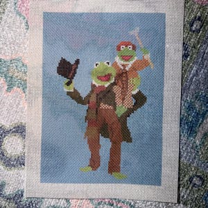 May include: A needlepoint canvas featuring two green frog characters against a light blue background. One frog wears a brown coat and scarf while holding a top hat, and the other frog sits on its shoulders wearing a brown jacket and holding a cane.