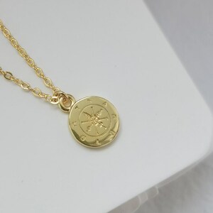 Könnte beinhalten: Eine goldfarbene Halskette mit einem Kompass-Anhänger. Der Anhänger ist eine kleine, runde Scheibe mit einem Kompassrosen-Design. Die Kette ist filigran und die Halskette wird auf einer weißen Oberfläche präsentiert.
