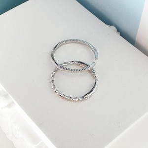 Puede incluir: Dos anillos de plata, uno con un diseño trenzado y el otro con una banda texturizada, están apilados uno encima del otro.