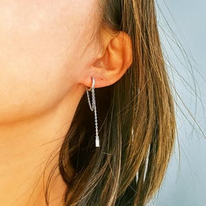 Könnte beinhalten: Ein silberner Creolenohrring mit einer baumelnden Kette und einem kleinen Diamanten-Akzent. Der Ohrring wird am linken Ohr einer Person mit langen braunen Haaren getragen.