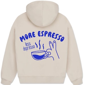 Könnte beinhalten: Beiger Kapuzenpullover mit blauem Aufdruck. Der Aufdruck zeigt den Text "MORE ESPRESSO less depresso", eine Kaffeetasse und eine Handillustration. Der Hoodie hat eine Kapuze und lange Ärmel. Das Design befindet sich auf der Rückseite.