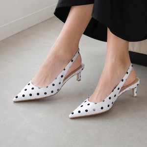 May include: White slingback kitten heels featuring a black polka dot pattern, a pointed toe, and a thin heel. The shoes have an adjustable strap at the back and a smooth finish, providing a classic look with a low heel height for everyday wear.