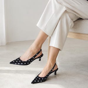 May include: Black slingback pumps featuring a white polka dot pattern, a pointed toe, and a low kitten heel. The shoes have an adjustable strap that wraps around the back of the heel, providing a secure fit while maintaining a sleek, low-profile silhouette.