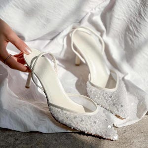 May include: A pair of white bridal heels with a delicate strap and a pointed toe. The heels are covered in sparkling sequins and beads.