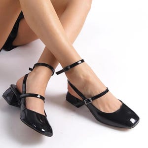 May include: Black patent leather Mary Jane style shoes with a square toe and a low block heel. The shoes have an ankle strap with a buckle closure and a strap across the top of the foot.