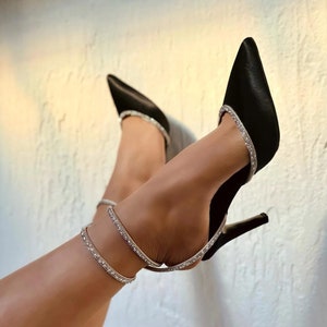 May include: Black satin high heel shoes with a rhinestone ankle strap. The shoes have a pointed toe and a thin stiletto heel.
