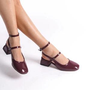 May include: Burgundy-colored, patent leather shoes with a square toe and a block heel. The shoes have an ankle strap and a strap across the top of the foot. The shoes are approximately 2 inches high.
