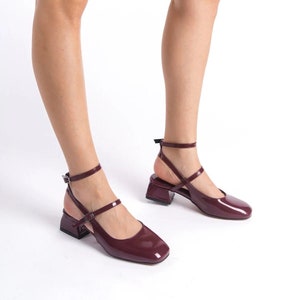 May include: Burgundy-colored, square-toe pumps with ankle straps and block heels. The shoes have a glossy finish and a modern design. The heels are approximately 2 inches high. The shoes are displayed on a white background.