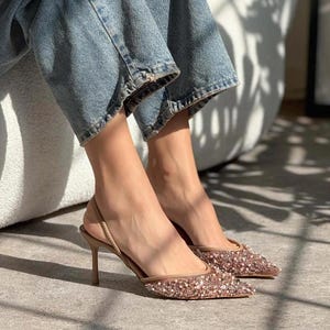 May include: A pair of nude-colored high-heeled shoes with a slingback strap. The shoes are covered in sparkly sequins and have a pointed toe.