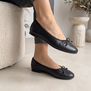 May include: A pair of black leather ballet flats featuring a rounded toe, a low heel, and a small decorative bow detail on the front. The shoes have a simple slip-on design with a thin trim along the opening and a smooth, matte finish throughout.