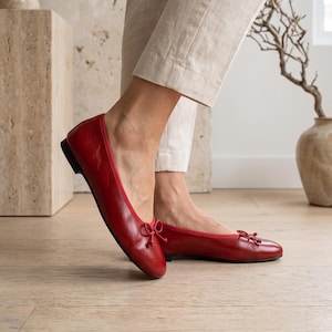 May include: A pair of red leather ballet flats featuring a rounded toe, a thin red trim along the collar, and a small decorative bow at the front. The shoes have a low, flat black heel and a smooth, polished finish, worn with light beige trousers.