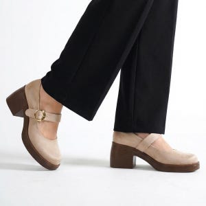 Pode incluir: Sapatos bege estilo Mary Jane com salto grosso castanho e sola plataforma. Os sapatos têm uma tira na parte superior do pé com uma fivela dourada. Os sapatos são usados com calças pretas. Altura do salto: aprox. 7,6 cm.