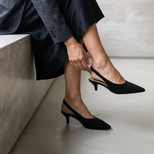 Könnte beinhalten: Schwarze Slingback-Heels mit spitzer Zehenpartie und kleinem Absatz. Die Schuhe haben einen Riemen um den Knöchel. Die Absätze sind etwa 5 cm hoch. Hergestellt aus einem weichen Material.