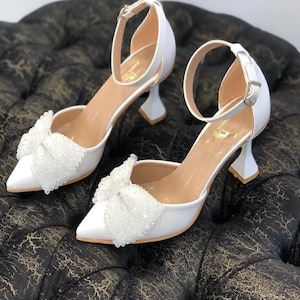 Puede incluir: Zapatos de novia blancos con un delicado detalle de lazo y una correa de tobillo. Los zapatos tienen una punta puntiaguda y un tacón de bloque bajo.