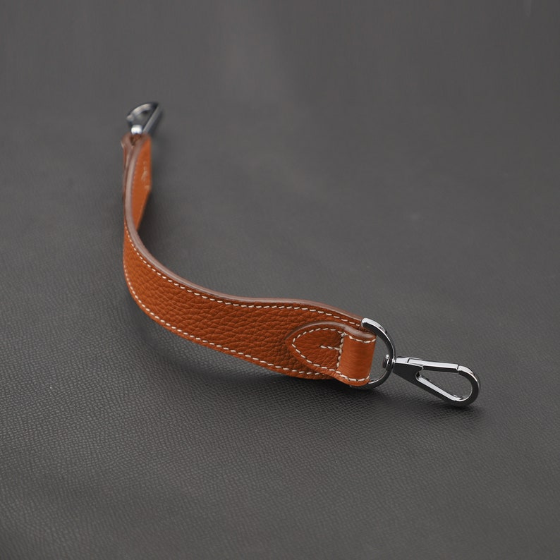 May include: A brown leather strap with a silver metal hook. The strap is stitched with white thread and has a slight curve.