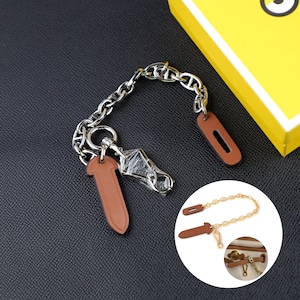 Op de afbeelding: Een zilverkleurige metalen ketting met twee bruine leren lipjes aan de uiteinden en een centrale zilverkleurige metalen sluiting. De ketting wordt getoond naast een cirkelvormige inzet met een goudkleurige versie van de ketting en een detail.