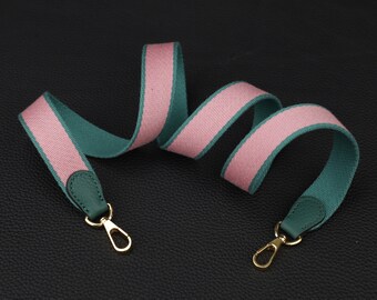 May include: A pink and teal striped bag strap with gold-colored clasps and dark green leather accents. The strap is made of a woven fabric and is designed to be attached to a handbag or purse.