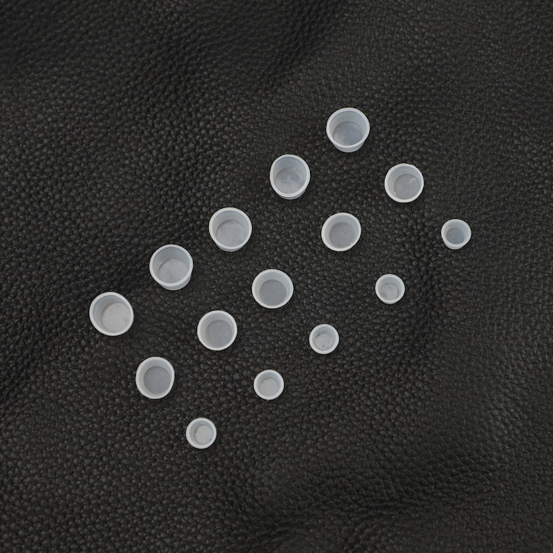 May include: A collection of 15 white plastic ink cups of various sizes, arranged in a triangular pattern on a black leather surface.