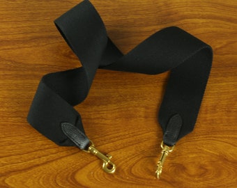 May include: Black fabric strap with gold metal clasps. The strap is folded in half and laid on a wooden surface.