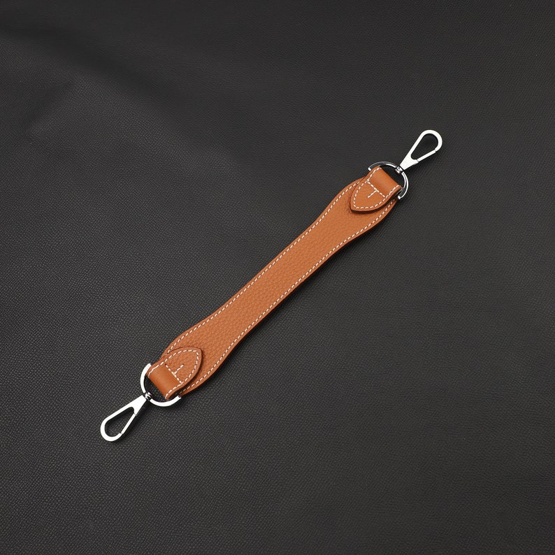 May include: A brown leather strap with two silver hooks. The strap is stitched with white thread.