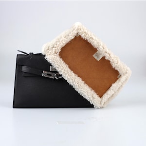 Peut inclure: Un sac à main en cuir noir avec une boucle et une poignée argentées est partiellement recouvert d'un article rectangulaire de couleur beige avec une bordure duveteuse de couleur crème. Le sac à main est de forme trapézoïdale structurée.