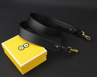 May include: A black leather camera strap with gold hardware and two adjustable loops. The strap is lying on a yellow box with a black and white eye design.