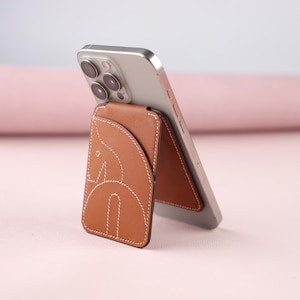 Può includere: Un supporto per telefono in pelle marrone con un design cucito bianco, che tiene uno smartphone argentato. Il supporto presenta un design astratto unico ed è appoggiato al telefono. Lo sfondo è rosa tenue.