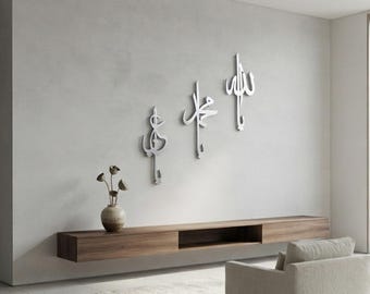 Puede incluir: Tres piezas de arte mural de metal plateado con caligrafía árabe vertical montadas en una pared gris claro sobre un estante flotante de madera horizontal largo que sostiene un pequeño jarrón de cerámica redondo con tallos florales secos.