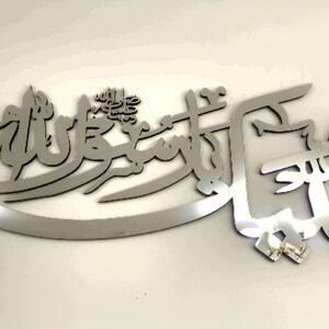 May include: A silver metal wall hanging with Arabic calligraphy. The script is in a flowing design, mounted on a white wall. This decorative artwork is suitable for home decor and measures approximately 50 cm in length.