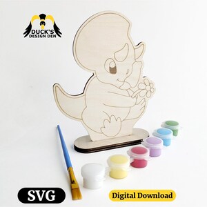 Puede incluir: Una figura de triceratops de madera sosteniendo una flor, lista para pintar. Incluye un pincel, botes de pintura de varios colores y el texto "SVG" y "Digital Download". El logotipo de Duck's Design Den está en la parte superior izquierda.