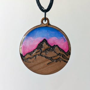 May include: Round wooden pendant with a mountain scene. The pendant features a mountain silhouette etched in dark brown, set against a backdrop of pink and blue resin. The pendant hangs from a black cord.