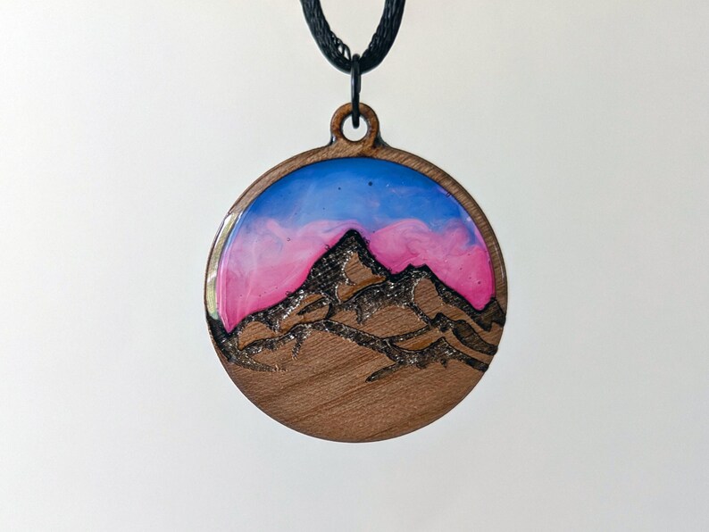 May include: Round wooden pendant with a mountain scene. The pendant features a mountain silhouette etched in dark brown, set against a backdrop of pink and blue resin. The pendant hangs from a black cord.