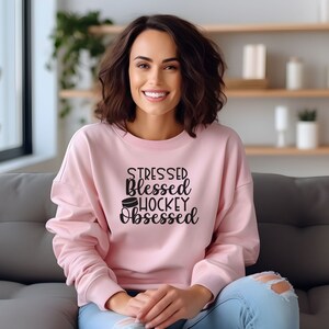K&ouml;nnte beinhalten: Ein rosa Sweatshirt mit dem Text "Stressed Blessed Hockey Obsessed" und einer Grafik eines Hockeyscheiben.