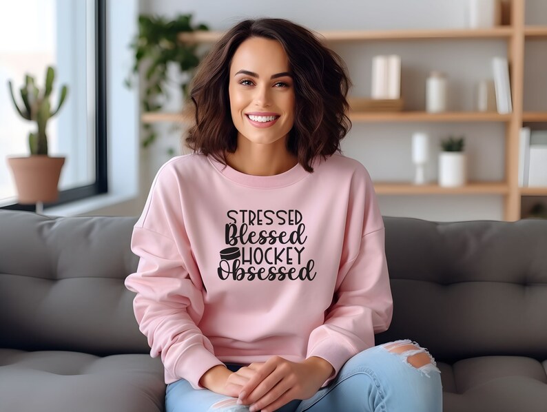 K&ouml;nnte beinhalten: Ein rosa Sweatshirt mit dem Text "Stressed Blessed Hockey Obsessed" und einer Grafik eines Hockeyscheiben.